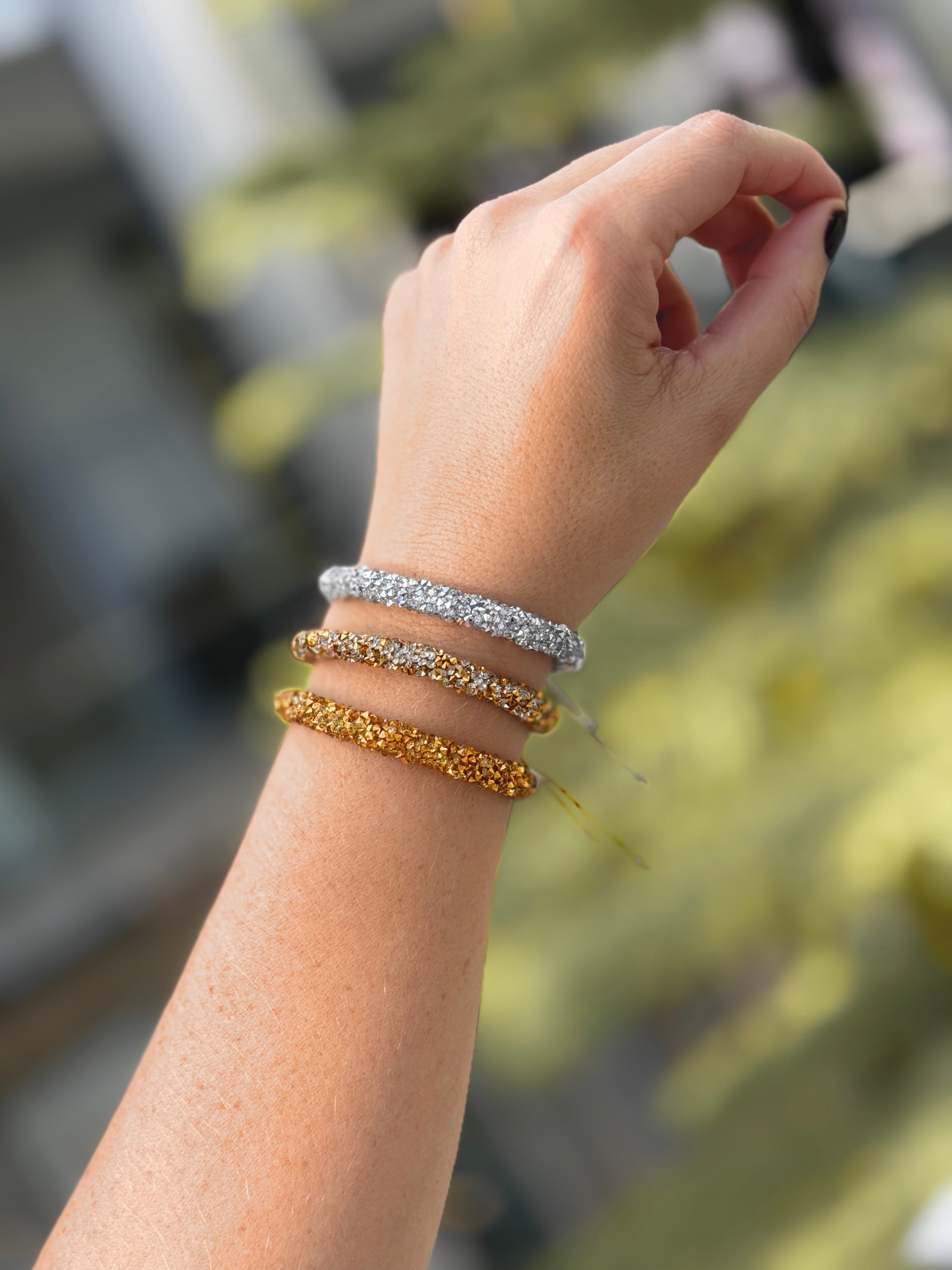 Three textured beaded bangles in silver, white, and gold tones stacked on a person's wrist, with a blurred outdoor background.