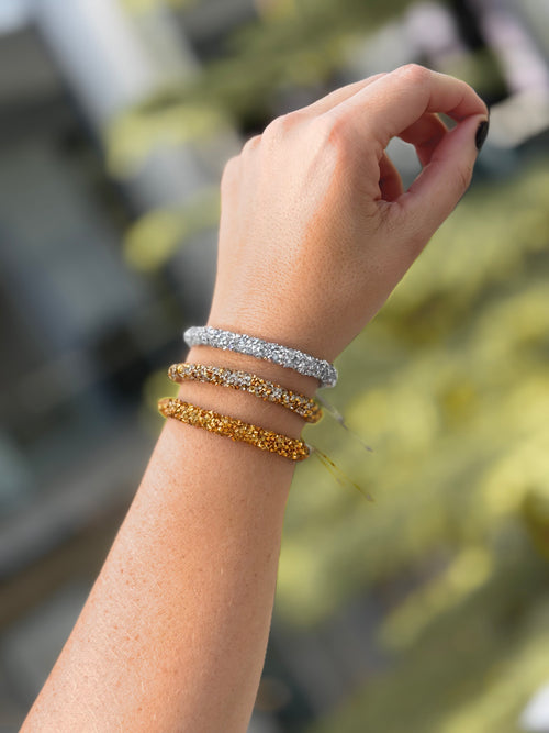 Three textured beaded bangles in silver, white, and gold tones stacked on a person's wrist, with a blurred outdoor background.