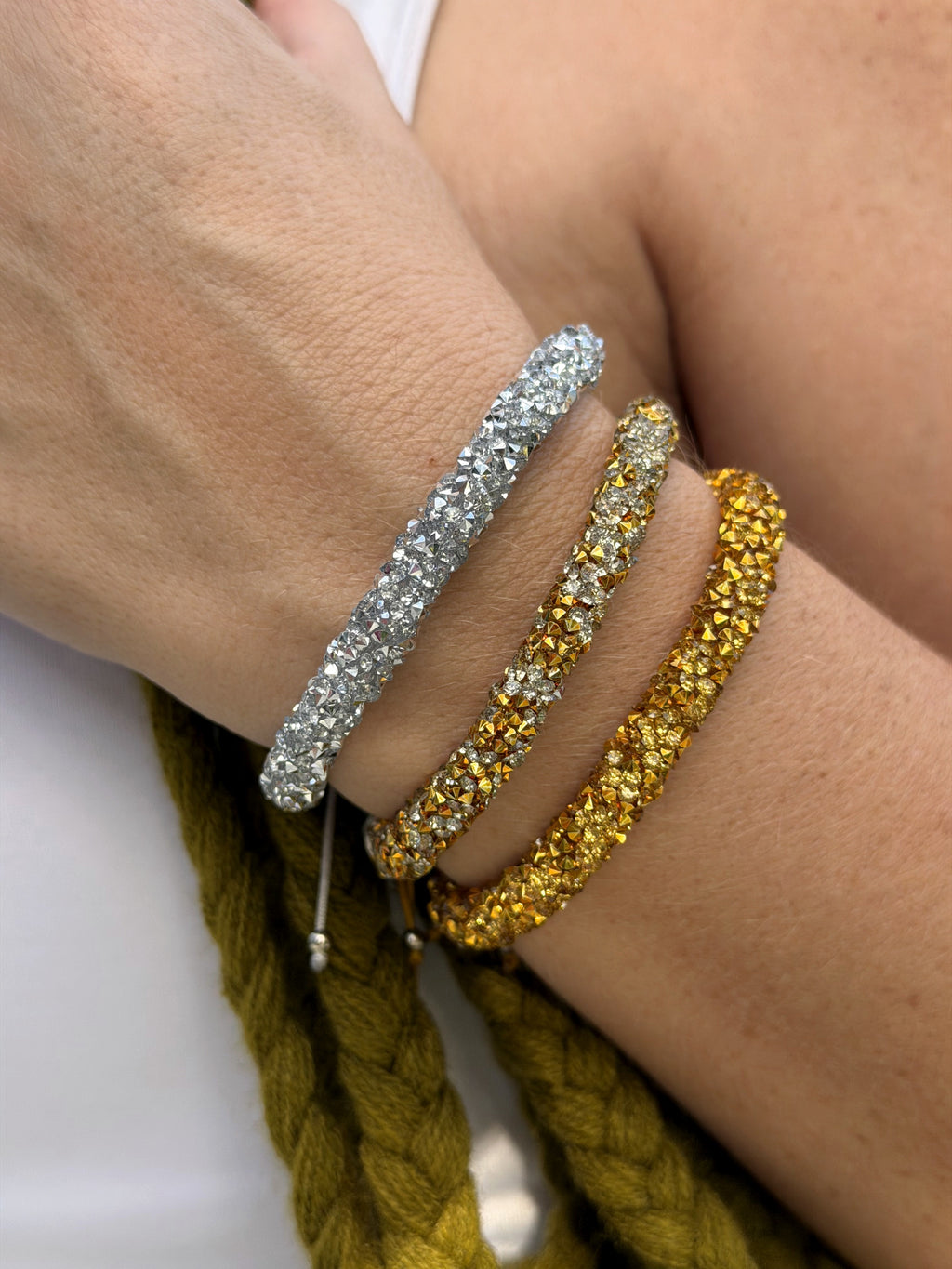 Three sparkling bangle bracelets worn on a person's wrist, featuring a silver band, a gold band with clear rhinestones, and a gold band with yellow rhinestones, shown against skin and a braided olive-green rope.