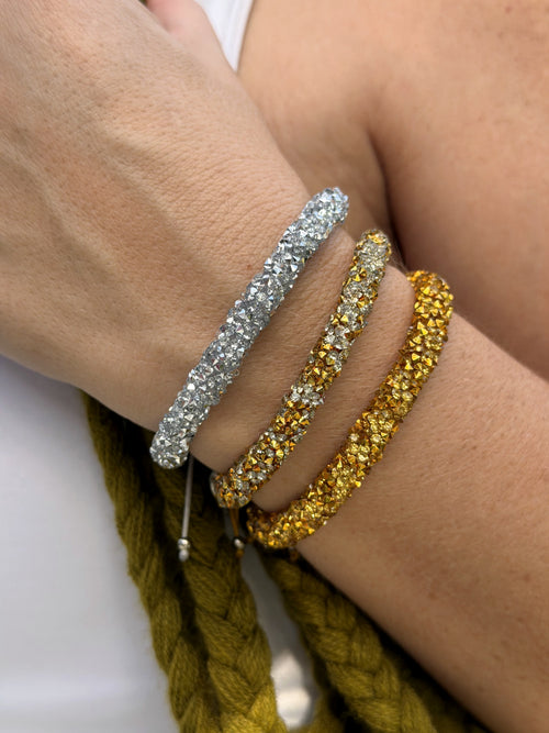 Three sparkling bangle bracelets worn on a person's wrist, featuring a silver band, a gold band with clear rhinestones, and a gold band with yellow rhinestones, shown against skin and a braided olive-green rope.