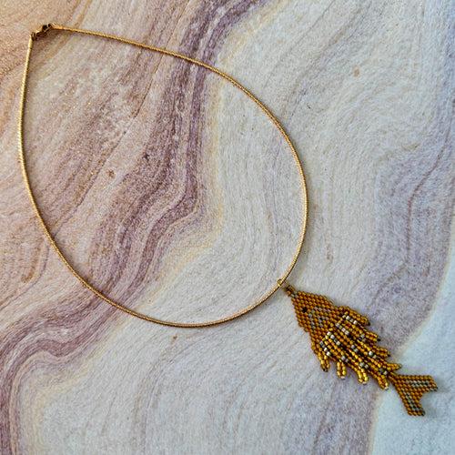 A gold-toned beaded necklace with a fish-shaped pendant, laid flat on a marbled stone surface with beige, gray, and purple veining.